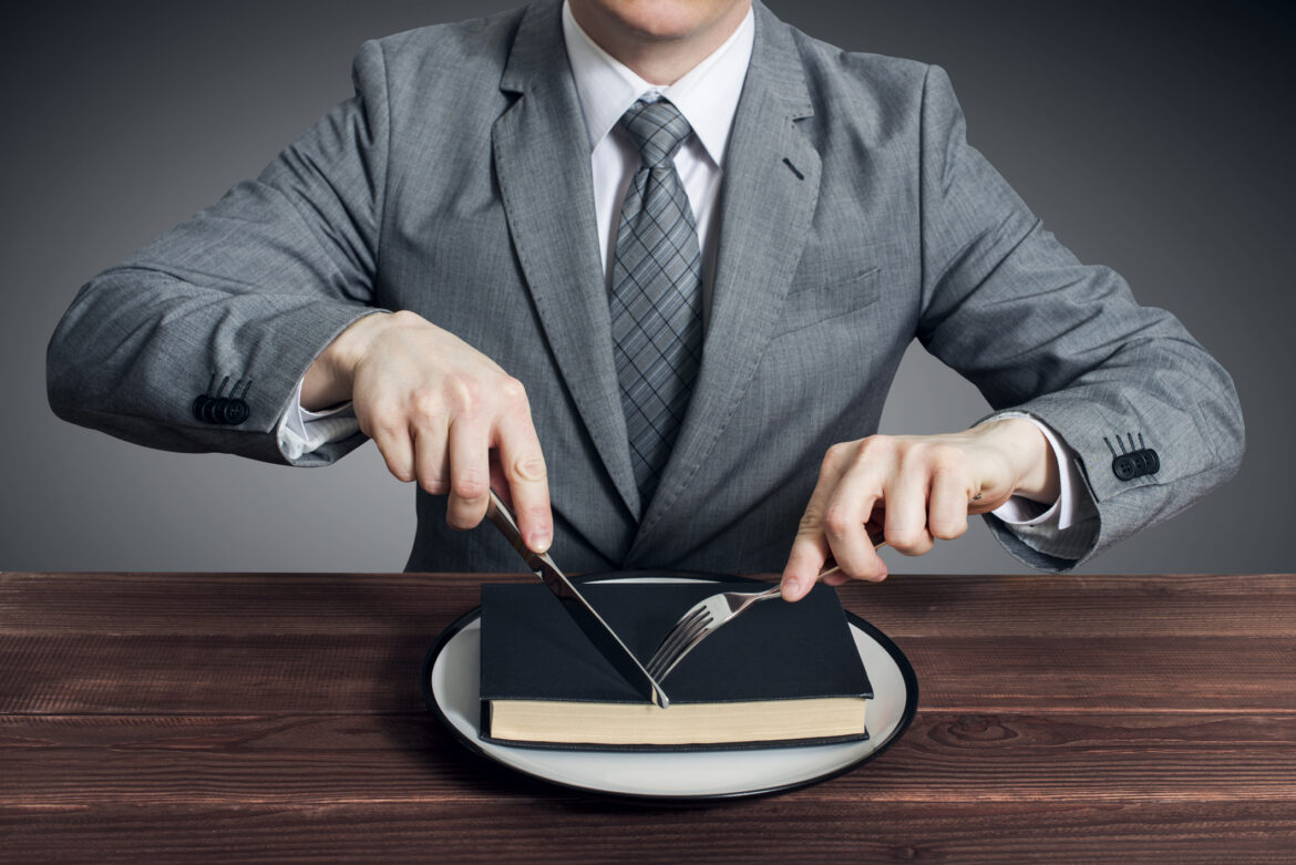 A businessman with a fork and knife eats a book on a plate
