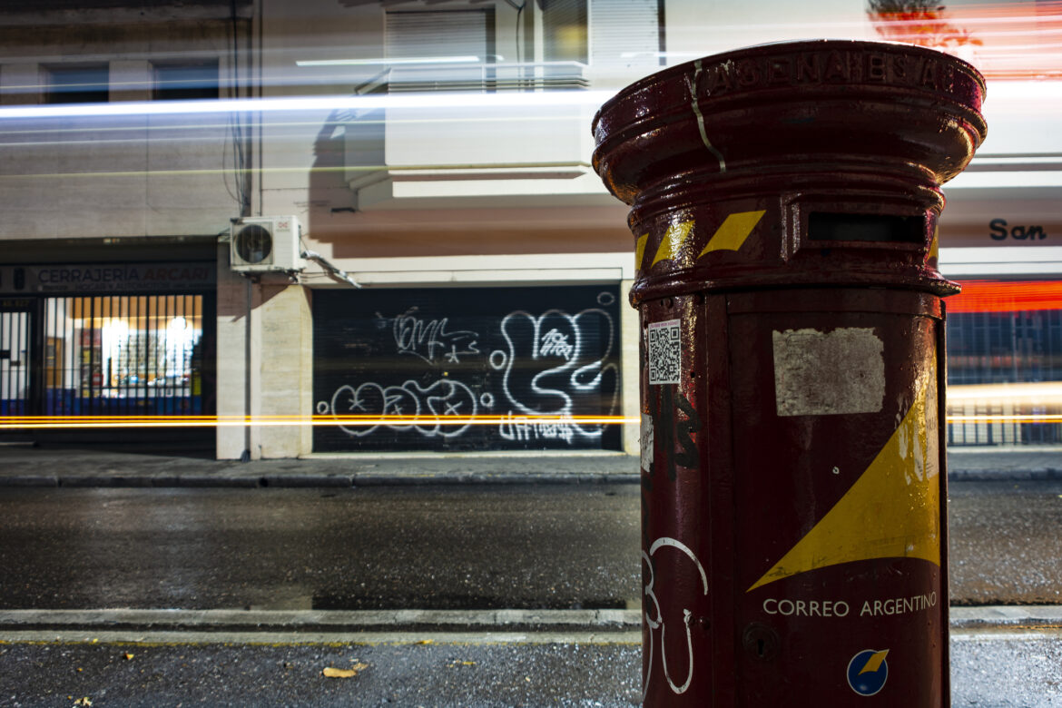 Urban scene nocturne, a red mailbox, in the right of the image, and bus lights passing for behind. Bright light and contrast photo in the public way of Rosario city, Santa Fe province, Argentina. On October 11, 2023.